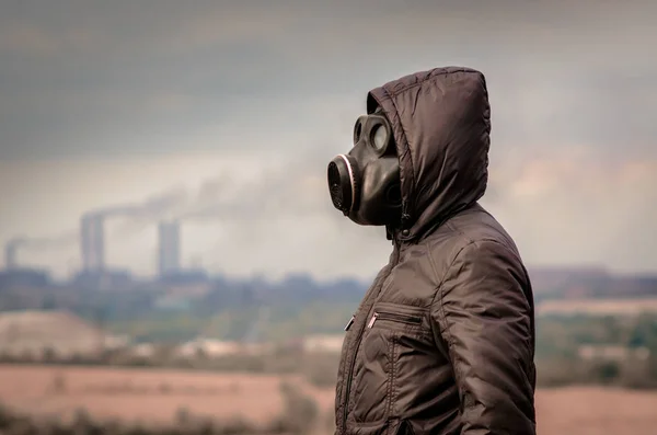 Hombre en una máscara de gas y una capucha en medio del humo de las tuberías de fábrica — Foto de Stock