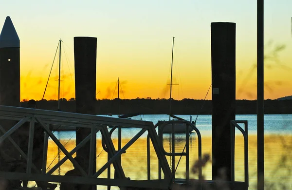 Gün batımı bir bay, bir feribot iskele üzerinde oturan balıkçı ile üzerinden. Su mavi ve altın. Koyda Yelkenli tekne palamarla ve tepeler mesafesindedir. Gökyüzü altındır.