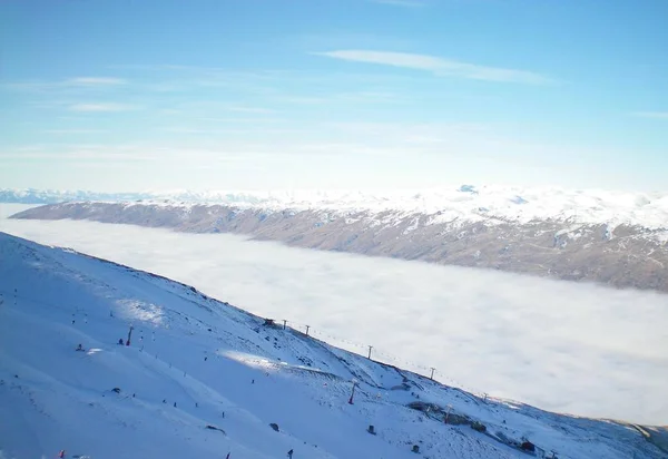 在满是云彩的山谷之上的滑雪场 Chairlifts 从云层中出来 滑雪者在山上 积雪覆盖的山脉在远处 天空是蓝色的 有白云 — 图库照片