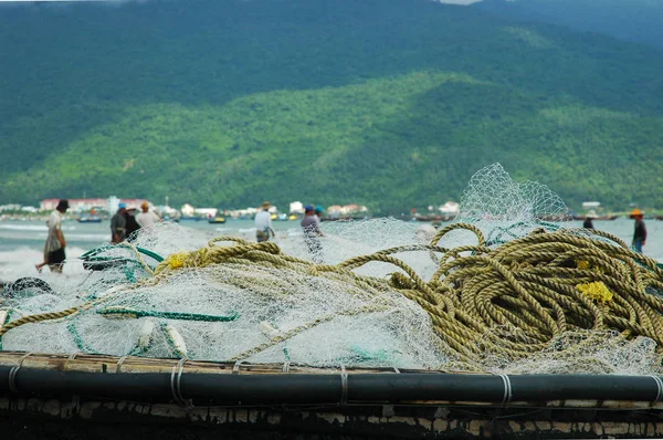 Fishing nets and rope rest on top of a traditioanl Vietnamese fishing boat. In the backgound, men stand on the shore, pulling in a fishing net. Forest covered hills are on the other side of the bay, with boats and houses on the shoreline.