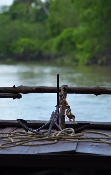 An anchor and rope rest on the back of a boat made from dark wood. A river flows past, boarded by the lush forest of the Mekong.
