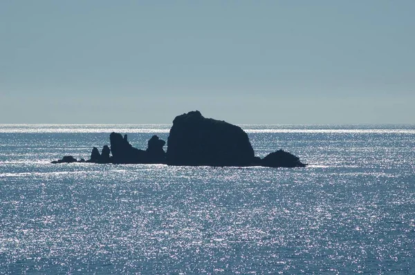 A group of rocks rising from the ocean. The rocks are black against the blue sky and water. The water is sparkling from the sunshine. The sky is clear, and the ocean calm.