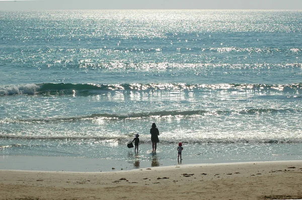 A mother is with her two young children at the beach. They stand at the water\'s edge. The water is calm, and sparkling under the sunshine. The sky is clear.