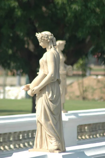 A statue in classic Greco-Roman style of a woman holding an urn. There is another statue in the background. Both are of white marble, and wear flowing drapes.