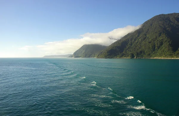 Wash from a boat spreading across the ocean. Forest covered hills run down to the shoreline. The sky is blue, with some clouds.