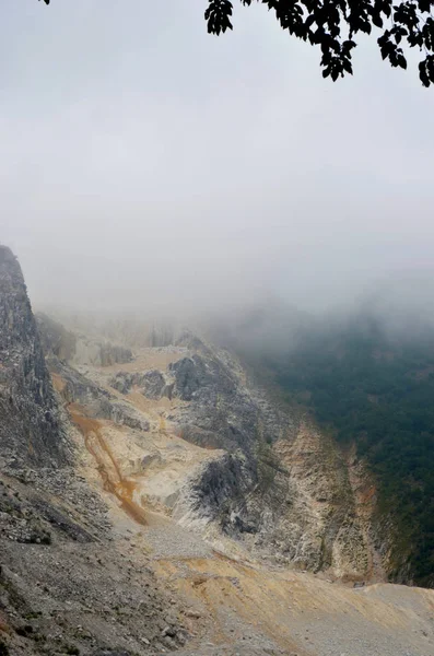 Mist covers a rock path through the mountains. The rocks are made of marble. Forest can be seen in the distance, and the leaves of a tree frame the view.