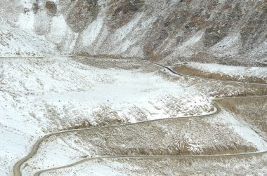 Kayalık bir yamacın üzerinden esen karla kaplı bir yol cüce gibidir. Beyaz bir araba ve yol tabelası zar zor görülebiliyor..