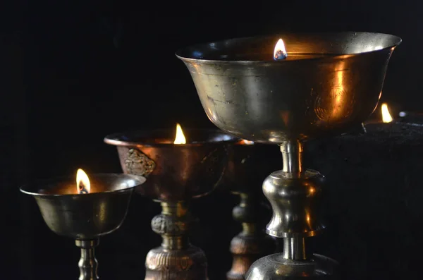 Old metal chalices are filled with oil and used as votive offerings. A flame is in each cup. Each cup is styled and decorated. The background is black.