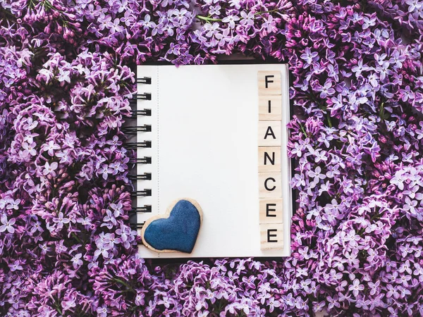 Heart-shaped cookie, covered with bright glaze, word FIANCEE, sketchbook with a blank page for Your inscription on the background flowers. Top view, close-up. Congratulations for relatives, loved ones