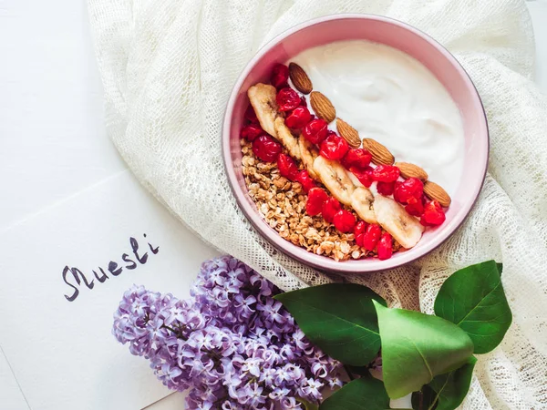 Healthy and delicious food. Bouquet of blooming lilacs, notebook with an inscription, berry smoothie, muesli with nuts, dry berries and pieces of banana in a pink plate on a stylish, white table