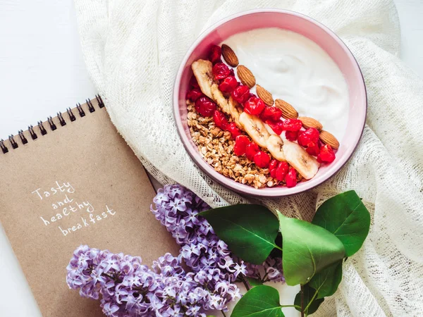 Healthy and delicious food. Bouquet of blooming lilacs, notebook with an inscription, berry smoothie, muesli with nuts, dry berries and pieces of banana in a pink plate on a stylish, white table