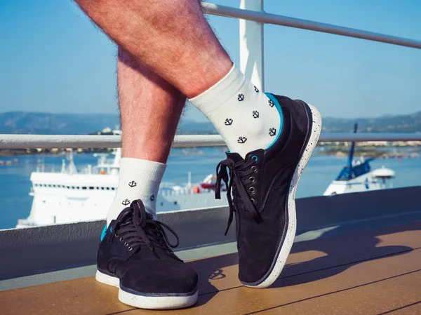 Men\'s feet in sport shoes and white socks with a sea pattern in the form of anchors on the open deck of a cruise liner against the background of the pool and the sea