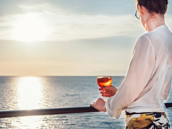 Young woman in glasses with a glass of beautiful, pink cocktail in hand on an open deck of a cruise ship on a background of sunset and sea waves
