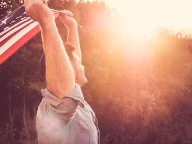 Çekici bir adam bir ABD bayrağı batan güneşin ışınları arka planı holding. Arkadan görüntüleyin. Tatil için hazırlanıyor