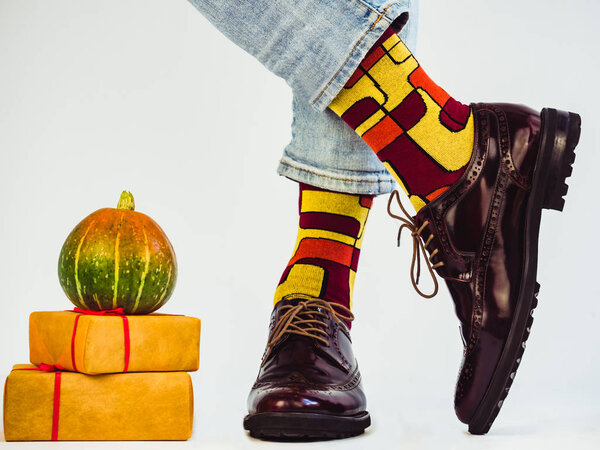 Beautiful pumpkins, gifts tied with a red ribbon, men's legs in stylish, vintage shoes and bright, multi-colored socks. White background, isolated, close-up. Holidays, fashion, elegance