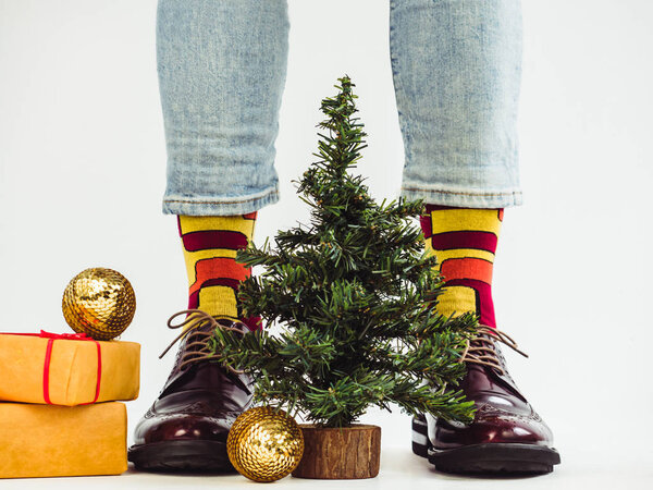Men's legs, funny socks, stylish shoes, gifts with a red ribbon and a Christmas tree. White background, isolated, close-up. Preparing for the holidays