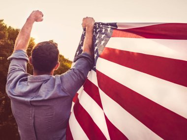 Rüzgar bize ayar güneş ışınlarının zemin karşı bayrak sallayarak tutan çekici bir adam. Arkadan, closeup görüntüleyin. Tatil için hazırlanıyor