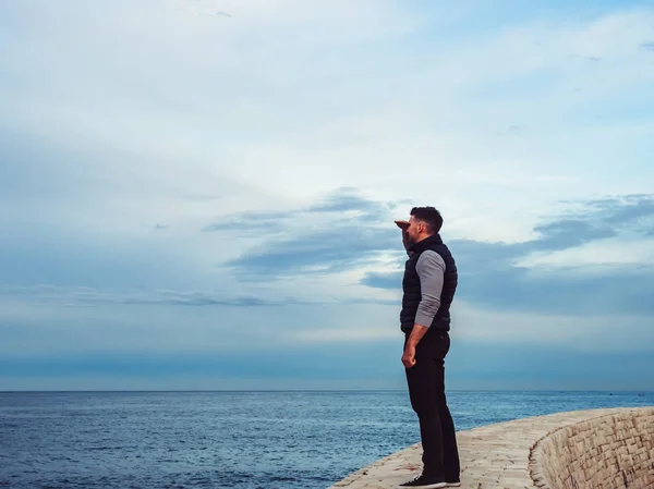 Fashionable, attractive man looking into the distance against the backdrop of sea waves and the coastline on a cloudy day