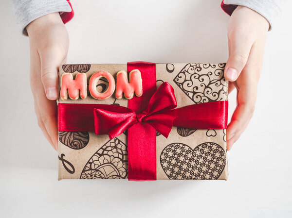 Children's hands, beautiful box with a gift, red ribbon and the word MOM on a white, isolated background. Top view, close-up. Preparing for the holidays