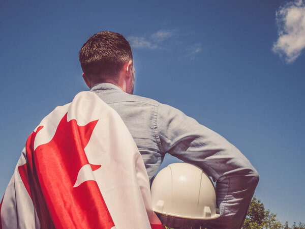 Young engineer, white hardhat and Canadian Flag in the park against the backdrop of green trees and the setting sun, looking into the distance. Close-up. Concept of labor and employment