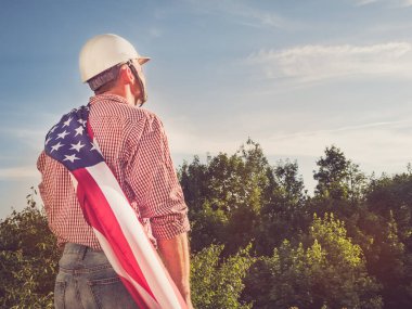 Genç mühendis, beyaz şapka ve parktaki Amerikan bayrağı yeşil ağaçların arka planına ve batan güneşe bakıyor. Yakın plan. Çalışma ve istihdam kavramı
