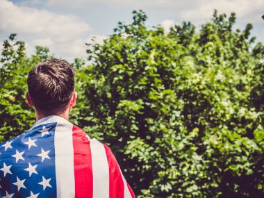 Açık ve güneşli bir günde mavi arka planda Amerikan Bayrağı tutan çekici bir adam. Arkadan bak, yakın plan. Ulusal tatil konsepti