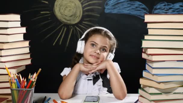 Modern skola koncept. Attraktiva flicka sitter vid ett skrivbord med högar av böcker. och en blackboard bakom henne. Skolflicka i hörlurar leenden. — Stockvideo