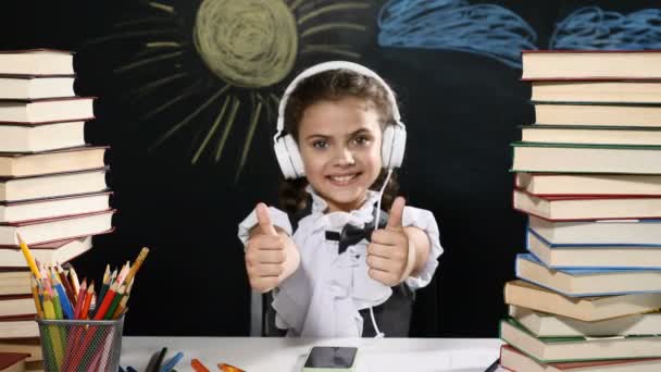 Concept d'école moderne. Jolie fille est assise à un bureau avec des tas de livres. et un tableau noir derrière elle. écolière positive dans les sourires casque. les pouces levés . — Video