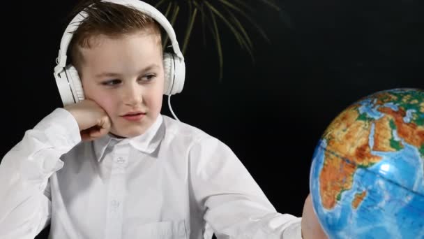 Schulkonzept. Netter Junge sitzt an einem Schreibtisch mit Globus darauf und einer Tafel hinter ihm. langweiliger Schüler dreht Globus um und zeigt auf einen zufälligen Ort — Stockvideo