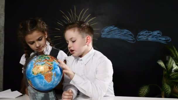 Concepto escolar. Los escolares se sientan en un escritorio jugando con un globo en él. Los niños se divierten apuntando a lugares al azar — Vídeos de Stock