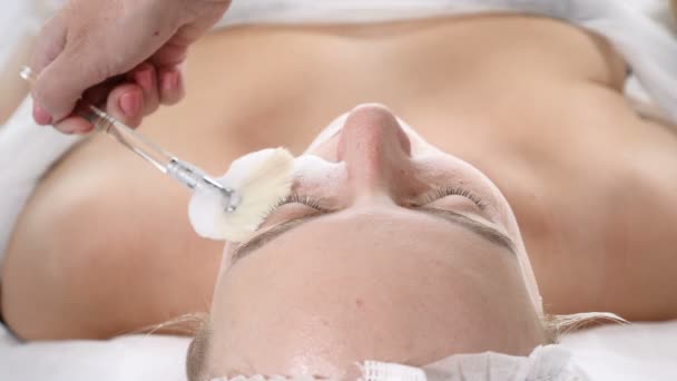 В клинике красоты. Молодая женщина проходит процедуру по уходу за лицом. Косметолог распыляет маску на женское лицо кистью . — стоковое видео