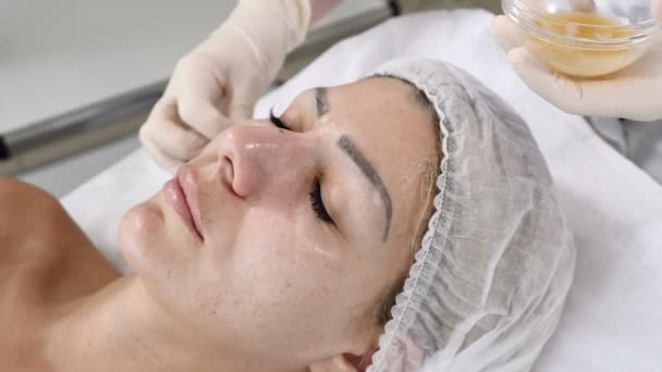 Pojęcie piękna. Młoda kobieta klienta pobiera piękno twarzy procedury. Renowacja twarz. Kosmetyczka używa wacikiem na pokrycie skóry twarzy z maską piękna — Wideo stockowe