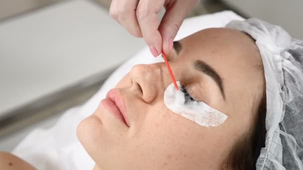 Mujer bonita joven recibiendo procedimientos en el salón de belleza. Joven hermosa mujer morena acostada en el sofá. Cosmetólogo usando espuma de limpieza facial en las pestañas — Vídeos de Stock