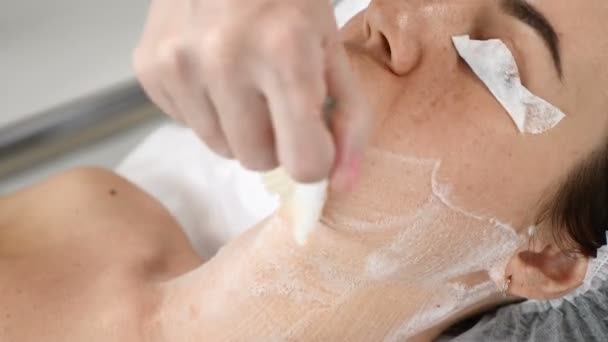 Young pretty woman getting procedures in beauty salon. Young beautiful dark-haired woman lying on couch. Cosmetologist using facial cleansing foam — Stock Video