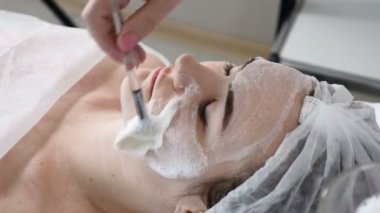 Genç kadın güzellik salonunda yordamlar almak güzel. Kanepede yatan genç güzel esmer kadın. Güzellik uzmanı yüzünde yüz temizleyici köpük uygulama