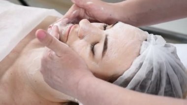 Genç kadın güzellik salonunda yordamlar almak güzel. Kanepede yatan genç güzel esmer kadın. Güzellik uzmanı yüz temizleyici köpük halinde kadın yüz ve boyun losyonları