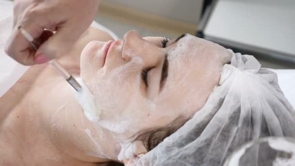 Mujer bonita joven recibiendo procedimientos en el salón de belleza. Joven hermosa mujer morena acostada en el sofá. esteticista aplicando espuma de limpieza facial en la cara — Vídeos de Stock