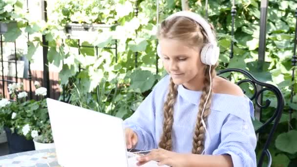 Portrét hezká dívka sedící v pouliční kavárně s notebookem na stole a sluchátka na hlavě. posloucháte hudbu, užívat si letních prázdnin. Volný čas před školou — Stock video