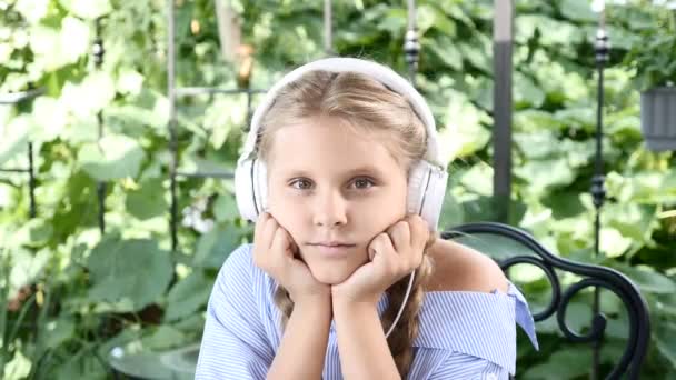 Portrait of pretty girl sitting in a street cafe with notebook on table and earphones on her head. listening to music, enjoying summer holidays. Free time before school — Stock Video