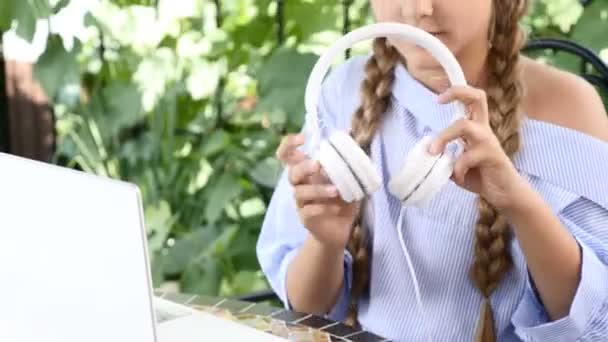 Videoretrato de una adorable chica sentada a la mesa con un cuaderno blanco, poniéndose auriculares — Vídeos de Stock