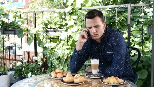 Business breakfast. Handsome young man in a street restaurant talking on mobile phone seriously. Variety of home baked treats are on table. man drinks coffee — Stock Video