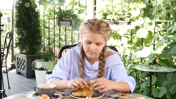 Porträt eines attraktiven kleinen Mädchens, das seine Freizeit in einem gemütlichen Café verbringt. junges Mädchen hält selbstgebackenes Croissant in der Hand und genießt den sonnigen Tag — Stockvideo