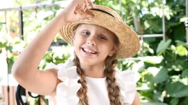 Маленька гарненька дівчинка розважається, коли її солом'яний капелюх посміхається. Стиль життя. Дівчина насолоджується літнім днем і гарним настроєм — стокове відео