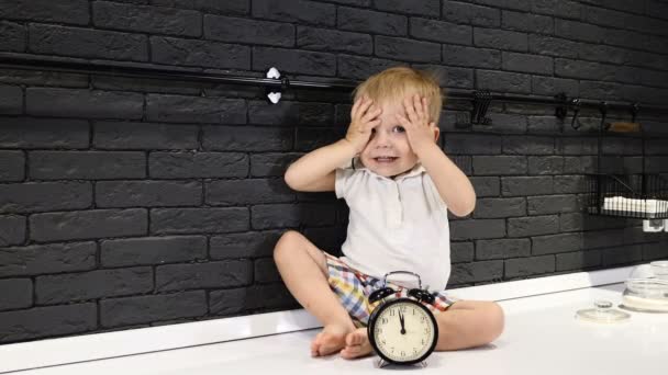 Porträt eines kleinen Jungen, der auf einem Küchentisch sitzt und neben ihm eine Uhr, die lächelnd die Augen schließt. Spaß haben. Fünf Minuten vor zwölf. 4k — Stockvideo