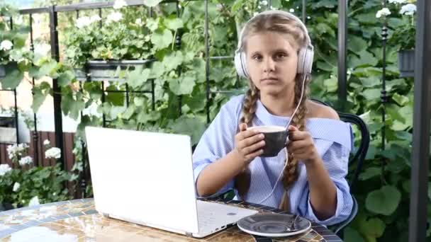 Porträt einer hübschen kleinen Dame, die mit einem Laptop beschäftigt ist und eine Tasse heiße Schokolade in der Hand hält. lächelnd in die Kamera. schönes Mädchen mit weißen Kopfhörern. 4k — Stockvideo