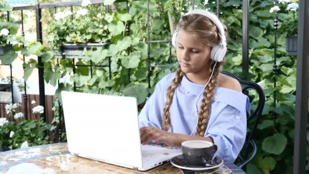 Junges hübsches Mädchen, das in einem Straßenrestaurant mit einem weißen Laptop auf dem Tisch sitzt. Lifestyle-Konzept. Schulmädchen hört Musik im Kaffeehaus mit Terrasse. 4k — Stockvideo