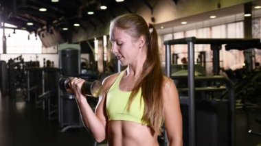 Elinde bir dumbbell ile atletik kadın spor salonunda. Aktif ve sportif genç kadın kameraya gülümseyen. Spor salonu arka plan. sağlıklı yaşam kavramı. 4k