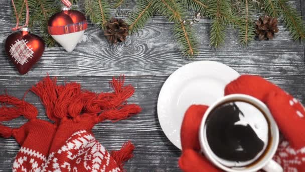 Buon Natale e felice anno nuovo concetto 2019 2020. Le mani in guanti lavorati a maglia rossi mettono una tazza di caffè caldo su uno sfondo di legno dove i simboli di anno nuovo sono messi. rami di abete, giocattoli di Natale — Video Stock