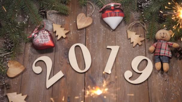 Bonne année 2019 concept. Vue de dessus sur les symboles en bois 2019 sur fond en bois décoré de branches de pin et de jouets faits main. Feu du Bengale tout autour. Mouvement lent — Video