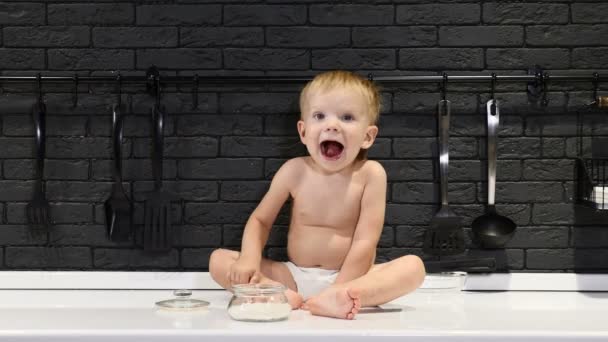 Lindo niño está en la mesa de la cocina comiendo azúcar sonriendo. Chico travieso probando dulces. 4k — Vídeo de stock